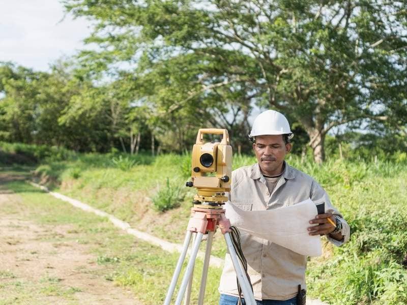 Professional land surveyor reviewing property survey plans with a total station at a residential site