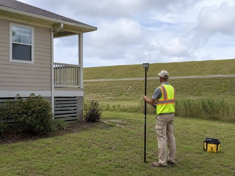 Land surveyor using GPS equipment to measure the elevation of a home located behind a grassy levee.